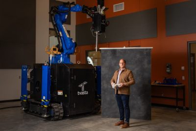 Andrew McCoy poses in front of a gray photo backdrop, next to a bright blue 3D concrete printer that is twice his height.